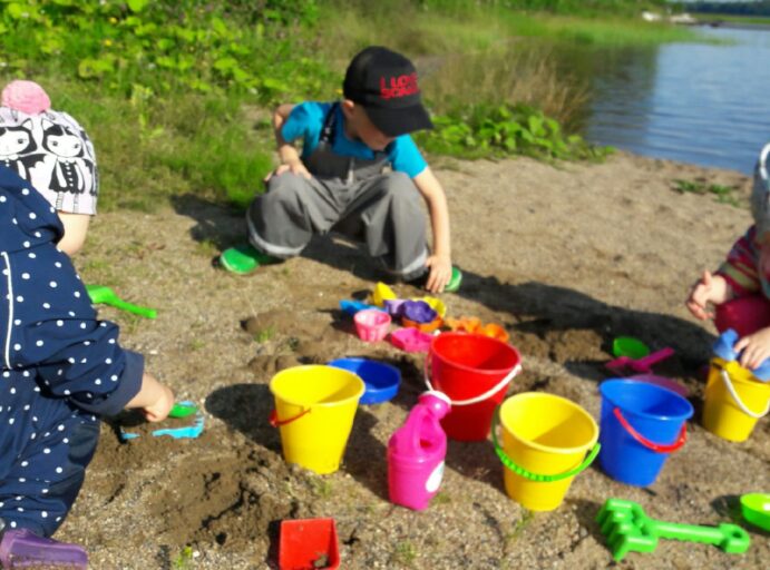 Kolme lasta leikkii rantahietikolla, hiekalla on erivärisiä hiekkasankoja ja hiekkaleluja.