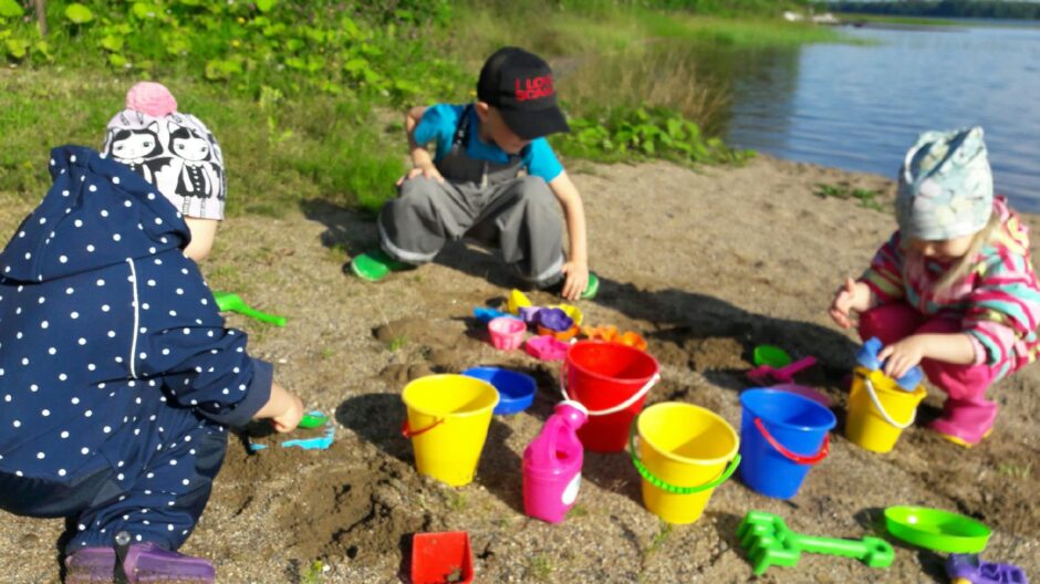 Kolme lasta leikkii rantahietikolla, hiekalla on erivärisiä hiekkasankoja ja hiekkaleluja.