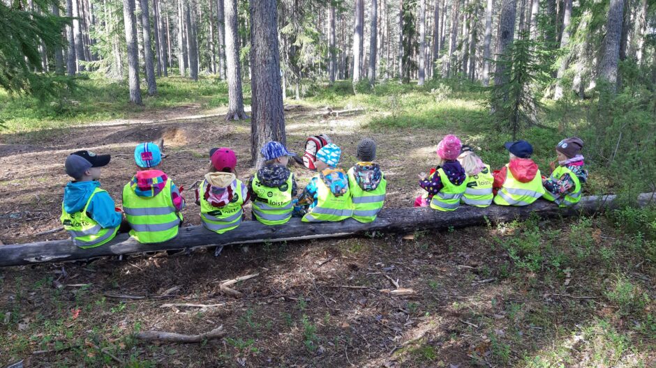 Aurinkoinen metsä, jossa kaatunut puunrunko etualalla. Puunrungolla istuu kymmenen metsäretkellä olevaa lasta selin, heillä on päällään keltaiset turvaliivit ja päässä värikkäitä pipoja ja lippiksiä.