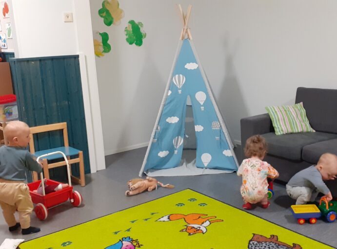Kolme lasta leikkii päiväkodin sisätiloissa, lattialla on matto, jossa on eläinten kuvia ja huoneessa on sininen kota, jossa ilmapallojen kuvia