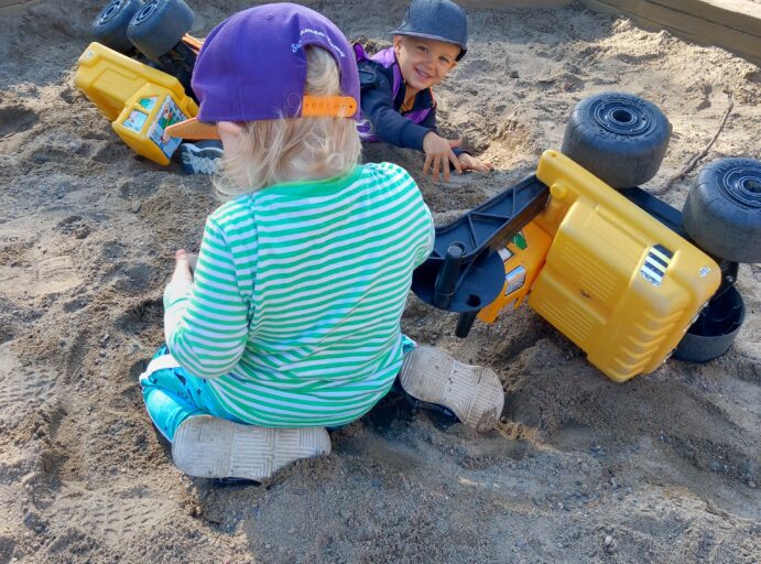 Kaksi lasta leikkii aurinkoisessa säässä hiekkalaatikolla kaivinkoneilla