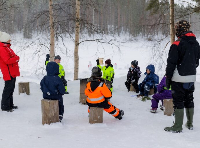 Lapset istuvat puupölkkyjen päällä lumisessa luonnosta kahden aikuisen kanssa.