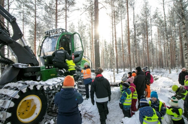 Talvinen metsä ja ryhmä lapsia tutustumassa metsäkoneeseen, jota aikuinen heille esittelee.