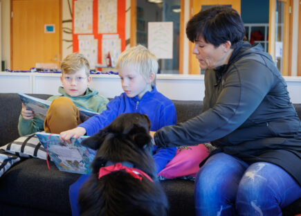 Lapset lukevat oppikirjaa sohvalla, ja opettaja sekä koira ovat opettamassa vieressä.