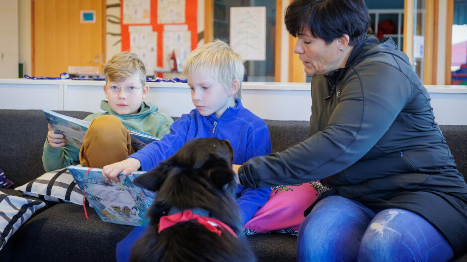 Lapset lukevat oppikirjaa sohvalla, ja opettaja sekä koira ovat opettamassa vieressä.