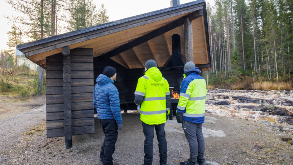 Rakennustarkastaja ja hankkeen osapuolen henkilöt katoksen katselmustilaisuudessa kosken rannalla.