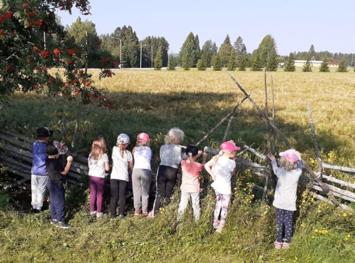 Kesäinen maalaismaisema, pelto ja taustalla puita, etualalla lapset selin nojaamassa heinäseipäistä tehtyyn aitaan.