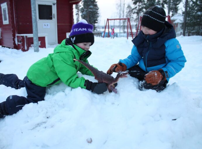 Kaksi lasta leikkii dinosauruksella lumessa.